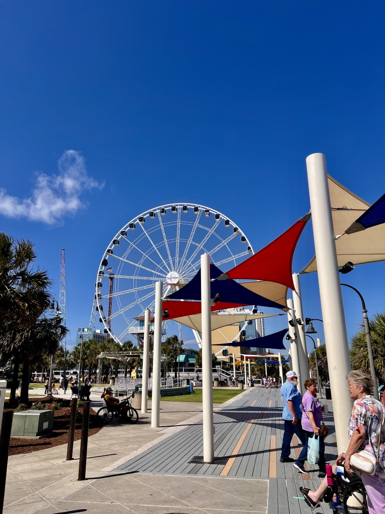 Myrtle Beach SkyWheel rising above the boardwalk with colorful shade sails and people walking nearby.