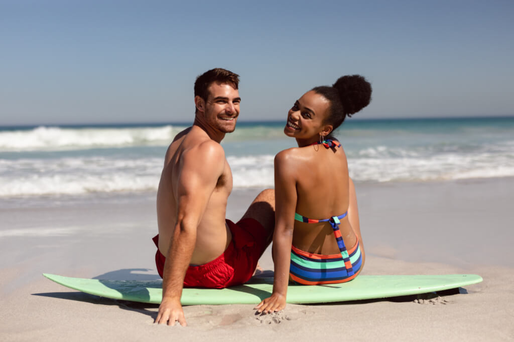 Young couple looking at camera while sitting on surfboard at beach in the sunshine