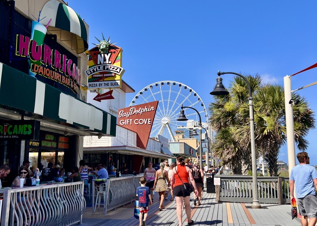 Crowds walking along the Myrtle Beach Boardwalk with the Gay Dolphin Gift Cove, Ferris wheel, and oceanfront shops.