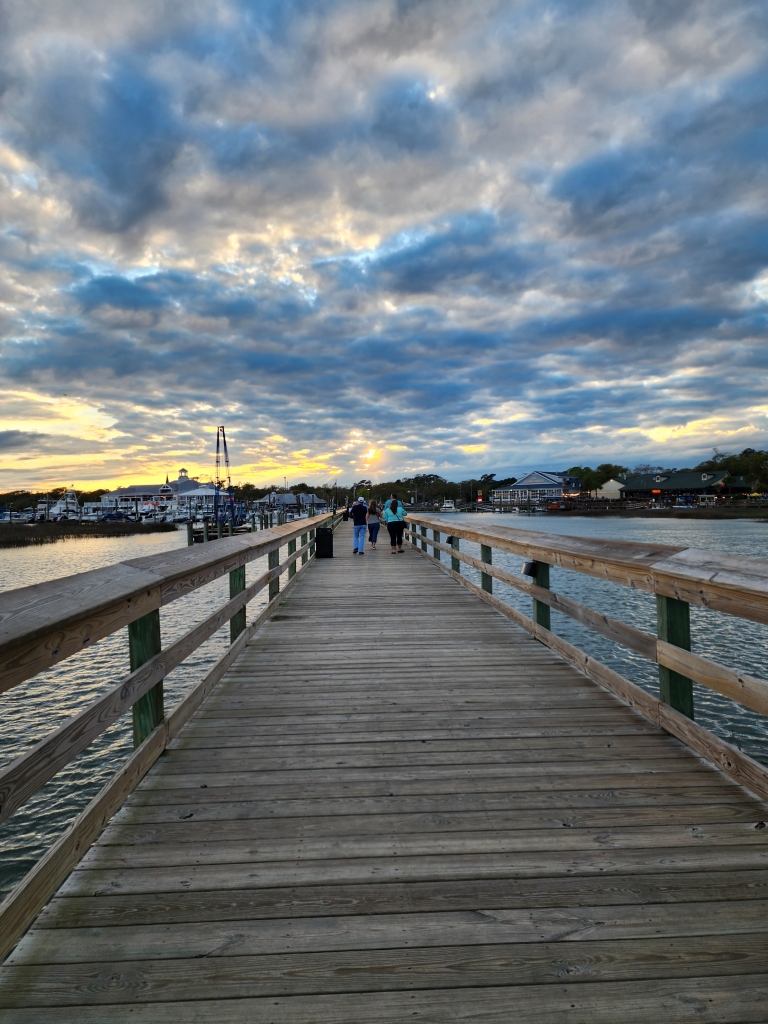 Wooden boardwalk over the water at sunset in Murrells Inlet, South Carolina, with people walking toward the waterfront restaurants