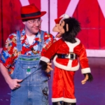 A ventriloquist performs on-stage with a puppet in a Christmas outfit.
