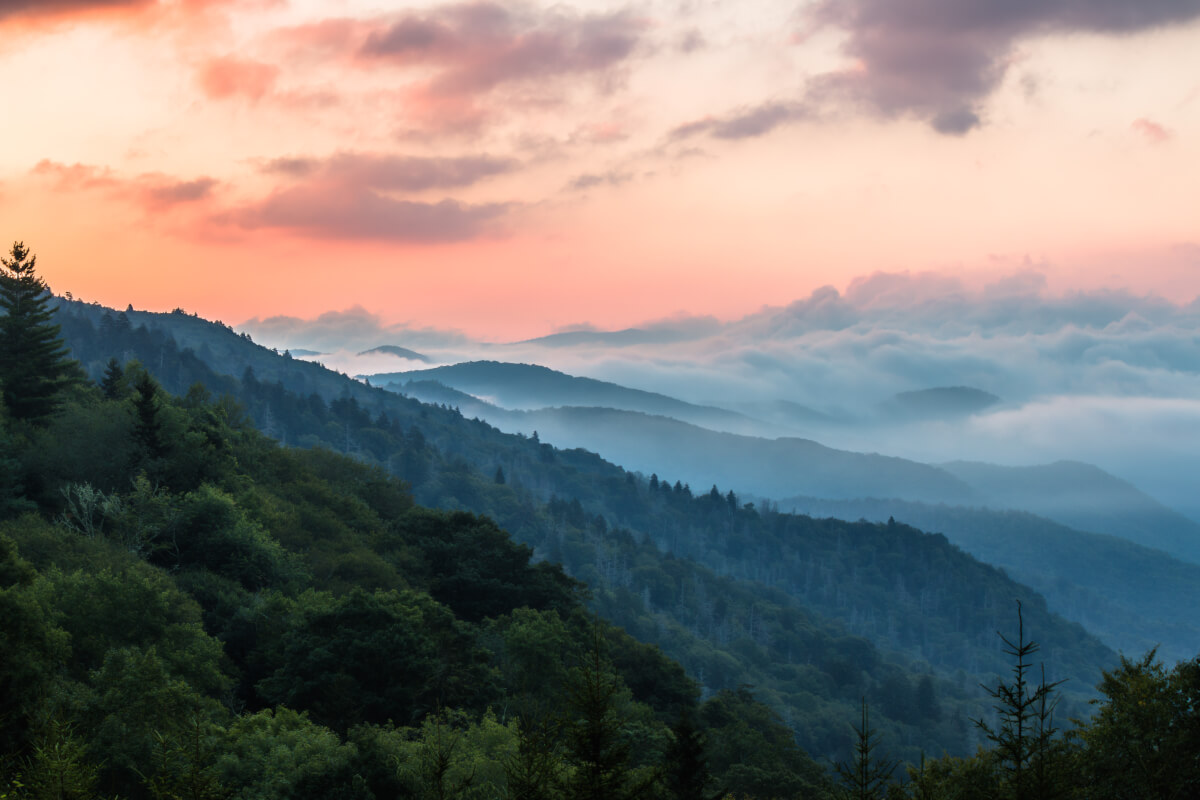 Sunrise over the Great Smoky Mountains with layered blue ridgelines, forested hills, and low clouds under a pink and orange sky.