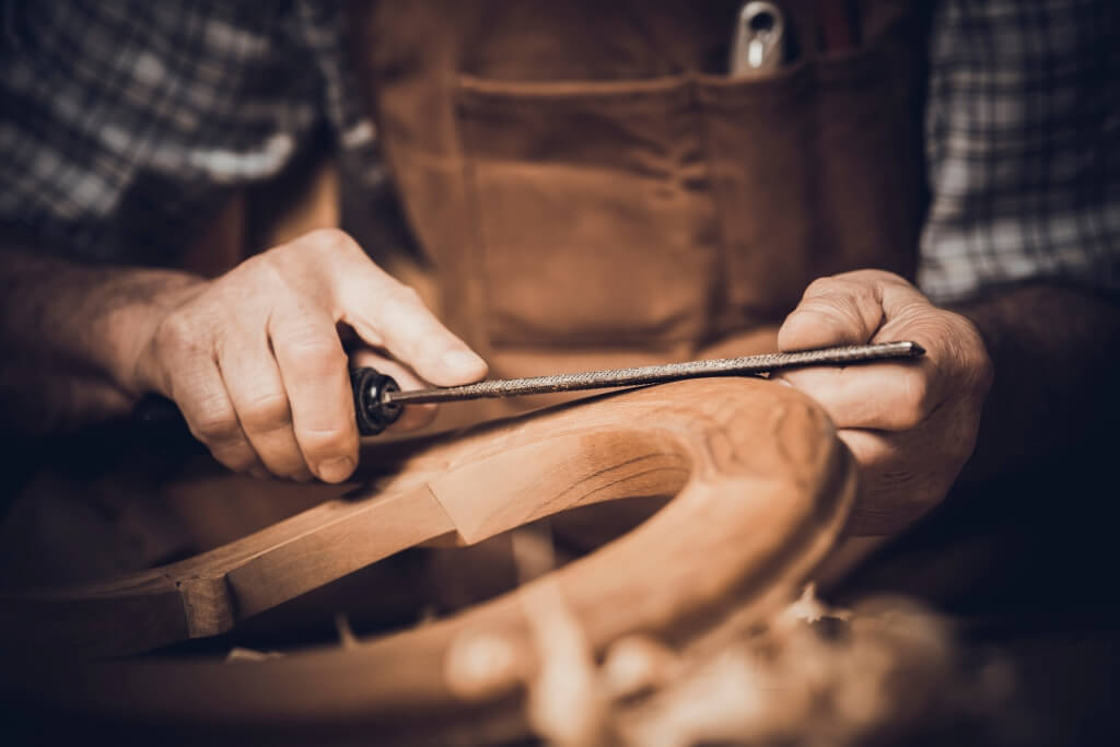 Close-up of a craftsperson shaping and smoothing a wooden piece by hand with a file in a workshop.