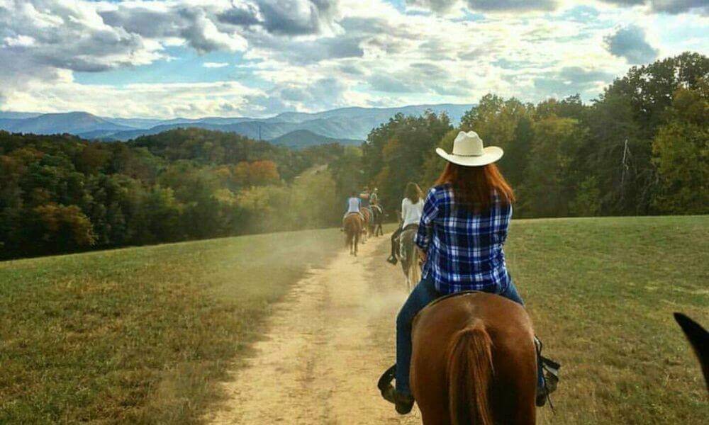 A person riding a horse on a mountain trail, surrounded by lush greenery and towering peaks.